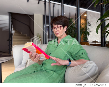 Middle-aged woman in green dress reading book on sofa. Knowledge, leisure, and personal time in modern lifestyle. 135252063