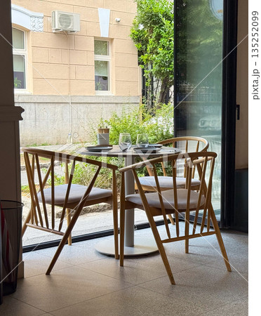 A wooden dining table with chairs and glassware stands by a window in a cafe. Hospitality, lifestyle, and leisure reflect urban dining culture and social atmosphere. A wooden dining table with chairs and glassware stands by a window in a cafe. Hospitality, lifestyle, and leisure reflect urban dining culture and social atmosphere. 135252099