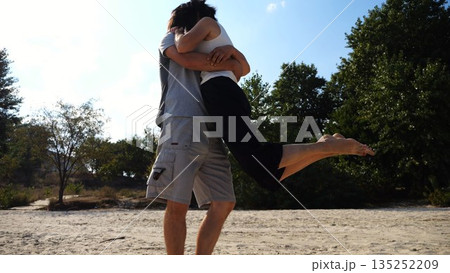 Happy couple having fun together on seashore at summer vacation. Man taking woman in air and turning around at coastline. Carefree lovers spending time at sandy beach. Concept of love. Slow motion 135252209