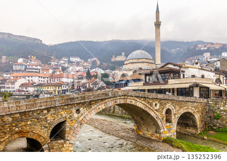 Stone arch bridge crossing a shallow Bistrica river with Ottoman Sinan Pasha mosque and tall minaret rising above dense hillside housing and traditional architecture on overcast day, Prizren, Kosovo Stone arch bridge crossing a shallow Bistrica river with Ottoman Sinan Pasha mosque and tall minaret rising above dense hillside housing and traditional architecture on overcast day, Prizren, Kosovo 135252396