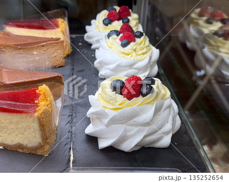Berry topped cream desserts and cheesecake slices behind cafe glass. Sweet indulgence, pastry craft, and modern bakery presentation through dessert display 135252654