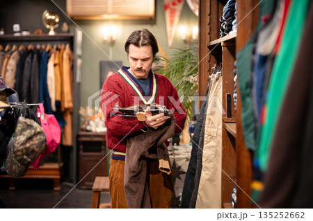 Hipster man conscious fashion customer choosing trendy velvet pants in second hand vintage store. Hipster man conscious fashion customer choosing trendy velvet pants in second hand vintage store. 135252662