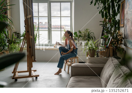 Relaxed woman artist looking at painting on easel while working at home loft studio with houseplants Relaxed woman artist looking at painting on easel while working at home loft studio with houseplants 135252671