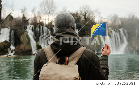 Traveler seen from behind holding Bosnia and Herzegovina flag while facing Kravica Waterfall. Exploration and reflection concept with wild nature, water scenery, and winter calm Traveler seen from behind holding Bosnia and Herzegovina flag while facing Kravica Waterfall. Exploration and reflection concept with wild nature, water scenery, and winter calm 135252981