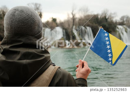 Traveler seen from behind holding Bosnia and Herzegovina flag while facing Kravica Waterfall. Exploration and reflection concept with wild nature, water scenery, and winter calm 135252983