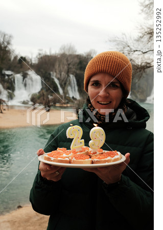 Smiling woman holding a cake in the form of sandwiches with red fish caviar with candles in front of Waterfall. Travel celebration concept with nature landscape, winter atmosphere, and joyful mood Smiling woman holding a cake in the form of sandwiches with red fish caviar with candles in front of Waterfall. Travel celebration concept with nature landscape, winter atmosphere, and joyful mood 135252992