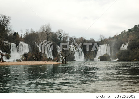 Wide cascading Kravica Waterfall surrounded by forest and turquoise river water in Bosnia and Herzegovina. Natural landscape with flowing streams, mist and calm atmosphere in early spring Wide cascading Kravica Waterfall surrounded by forest and turquoise river water in Bosnia and Herzegovina. Natural landscape with flowing streams, mist and calm atmosphere in early spring 135253005