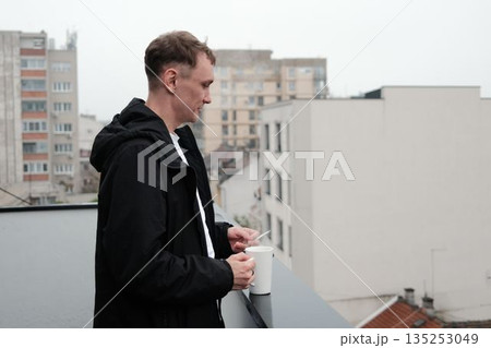 A man stands on a terrace holding a cup and a cigarette while observing the city. The scene suggests a slow morning concept 135253049