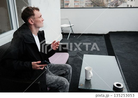 A man sits on a terrace smoking a cigarette and looking into the distance. The scene conveys a reflective morning concept 135253053