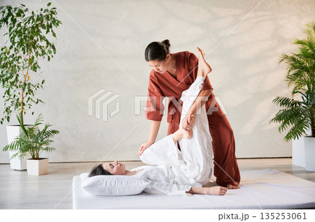 Masseuse making thai yoga massage. Woman therapist doing traditional massage treatment, stretching female patient's body. Client lying on mat, enjoying hand massage in calm atmosphere. 135253061