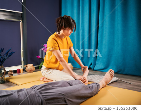 Masseuse making thai yoga massage. Woman therapist doing traditional massage treatment for male patient indoors. Client lying on mat, meditating, enjoying hand massage in calm atmosphere. Masseuse making thai yoga massage. Woman therapist doing traditional massage treatment for male patient indoors. Client lying on mat, meditating, enjoying hand massage in calm atmosphere. 135253080