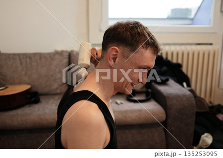 A man uses a massage gun on his neck while sitting on the floor. The scene conveys home relaxation concept A man uses a massage gun on his neck while sitting on the floor. The scene conveys home relaxation concept 135253095