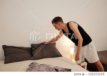 A man leans over the bed fixing and arranging pillows after waking up. The concept conveys tidiness, morning order, and establishing routine 135253229