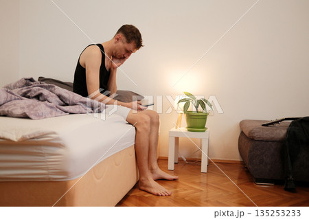 A man sits on the edge of the bed looking at his phone with a sleepy expression. The concept reflects a slow morning start and daily habit 135253233