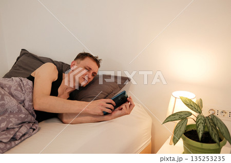 A man lies in bed holding a smartphone while a bedside lamp glows warmly beside him. The concept highlights early digital engagement and a soft morning mood 135253235