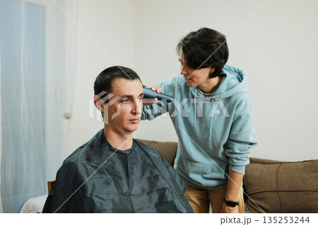 A woman carefully trims the sides of a man s hair during a home haircut. The scene highlights practical grooming and the idea of simple daily maintenance 135253244
