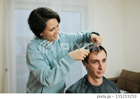 A woman trims a man s hair at home using scissors while he sits calmly. The scene presents everyday grooming and the concept of casual personal care A woman trims a man s hair at home using scissors while he sits calmly. The scene presents everyday grooming and the concept of casual personal care 135253250