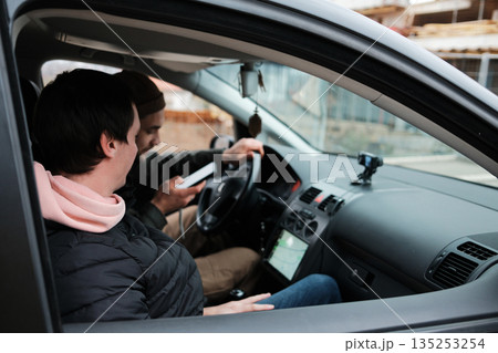 Two men sit inside a car while one checks information on a smartphone. The scene reflects navigation, planning, and road assistance Two men sit inside a car while one checks information on a smartphone. The scene reflects navigation, planning, and road assistance 135253254
