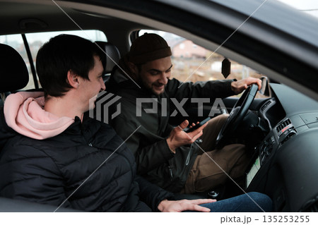 Two men sit inside a car while one checks information on a smartphone. The scene reflects navigation, planning, and road assistance Two men sit inside a car while one checks information on a smartphone. The scene reflects navigation, planning, and road assistance 135253255