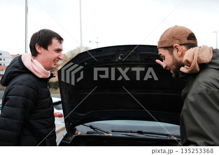 Two men stand by an open car hood discussing a mechanical issue. The moment reflects roadside support and friendly communication Two men stand by an open car hood discussing a mechanical issue. The moment reflects roadside support and friendly communication 135253368