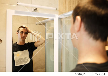 A man stands in front of a bathroom mirror and touches his head. The moment shows thought, routine, and personal control 135253381
