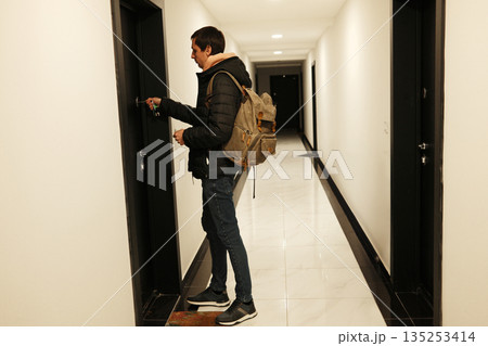 A man stands in a hallway with a backpack and places a key in the door lock. The image reflects everyday travel and the concept of returning home A man stands in a hallway with a backpack and places a key in the door lock. The image reflects everyday travel and the concept of returning home 135253414