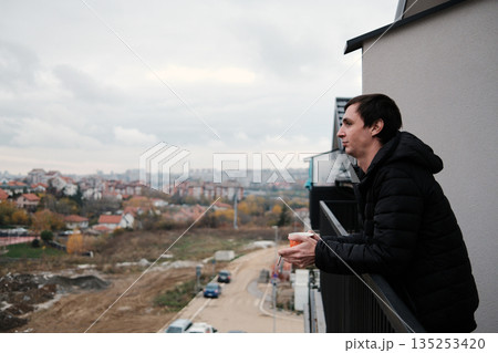 A man holds a cigarette and a cup while looking into the distance from a balcony. The scene conveys a calm break and the concept of personal time 135253420