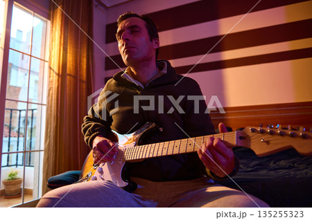 Relaxed Musician Playing Electric Guitar in Cozy Room With Sunset Light and Striped Wall 135255323