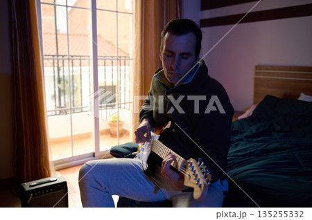Focused Musician Plays Electric Guitar in a Cozy Bedroom by Balcony Window During Evening Night 135255332