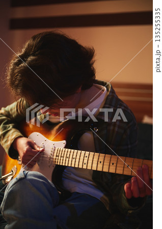 Young Man Playing Electric Guitar At Home In Warm Lighting, Casual Feel, Focused Expression Young Man Playing Electric Guitar At Home In Warm Lighting, Casual Feel, Focused Expression 135255335