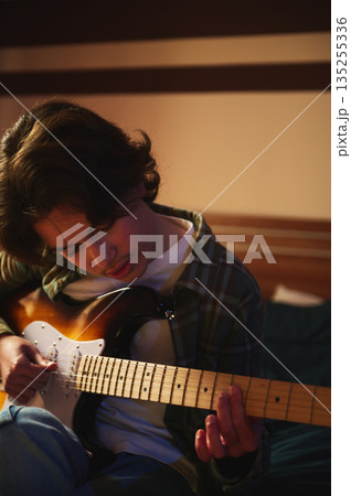 Young Man Plays Sunlit Electric Guitar on Bed in Cozy Home Setting Young Man Plays Sunlit Electric Guitar on Bed in Cozy Home Setting 135255336