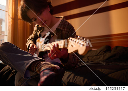 Young Man Strumming Electric Guitar On Bed During Golden Sunset At Home Young Man Strumming Electric Guitar On Bed During Golden Sunset At Home 135255337