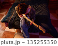 Young Person Strumming Electric Guitar on Bed in Cozy Bedroom with Warm Lighting 135255350