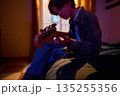 Young Man With Acoustic Guitar Sitting On Bed In Cozy Room Playing Music 135255356