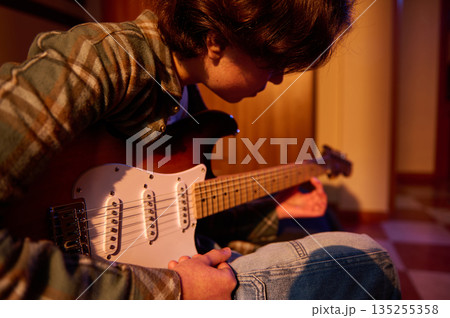 Young Musician Sits With Electric Guitar In Warm Cozy Room, Focused On Practice 135255358