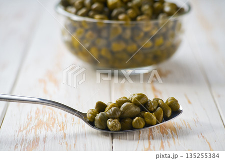 Metal spoon with pickled caper berries on a light kitchen table, selective focus. Metal spoon with pickled caper berries on a light kitchen table, selective focus. 135255384