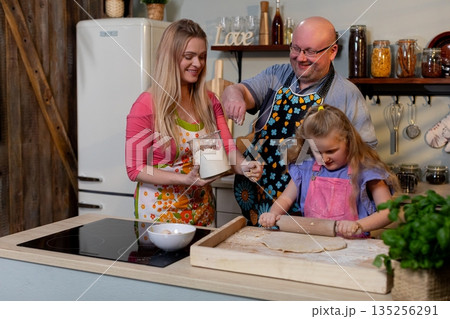 Parents and child tasting batter from mixing bowl, joyful exchange with spoon and bowl, cozy kitchen lighting and decorative jars enhance celebratory home cooking mood 135256291