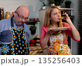 Woman sampling bite while partner watches, expressive tasting moment with plate and spoon, stove and spice jars in frame, casual recipe feedback and flavor evaluation 135256403
