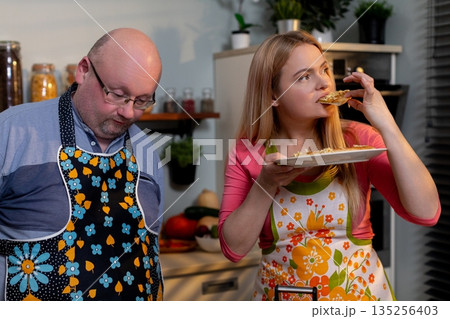 Woman sampling bite while partner watches, expressive tasting moment with plate and spoon, stove and spice jars in frame, casual recipe feedback and flavor evaluation 135256403