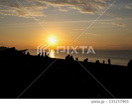 Silhouette of people enjoying the sunset on a beach, silhouetted figures against the stunning orange sky and calm sea with the natural light, creating a serene atmosphere Silhouette of people enjoying the sunset on a beach, silhouetted figures against the stunning orange sky and calm sea with the natural light, creating a serene atmosphere 135257903