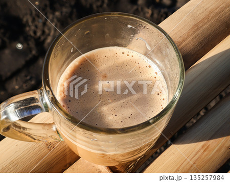 Frothy coffee in a clear glass mug on bamboo wood surface, casting shadows, perfect for a cozy morning drink presentation Frothy coffee in a clear glass mug on bamboo wood surface, casting shadows, perfect for a cozy morning drink presentation 135257984