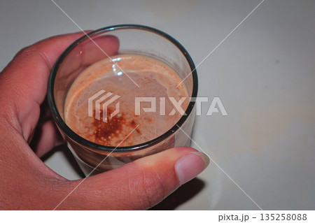 Hand holding glass of chocolate milk with cinnamon under natural light, showcasing homemade beverage for healthy lifestyle concept 135258088