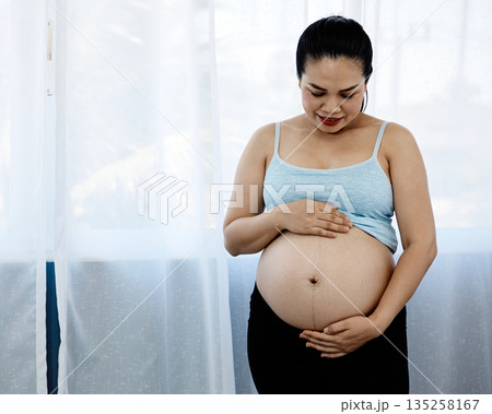 Asian pregnant woman standing indoors, gently holding her bare belly and smiling with love, representing prenatal bonding, maternal care, healthy pregnancy, and emotional connection to unborn baby 135258167