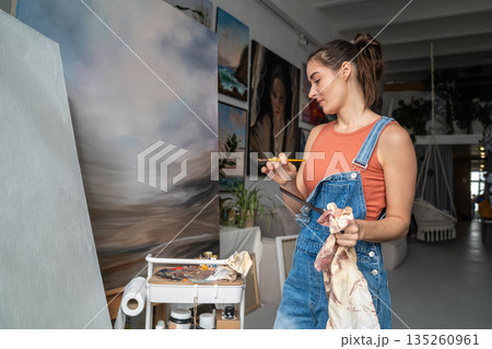 Smiling relaxed woman artist use tools and mediums for painting abstract picture on canvas in studio 135260961