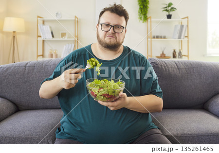 Unhappy man eating salad on sofa at home, struggling with healthy choices Unhappy man eating salad on sofa at home, struggling with healthy choices 135261465