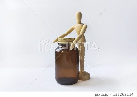 wooden mannequin stands against a white background next to a brown glass jar with gold lid. The mannequin appears to be interacting with the jar. The scene explores themes of health and well-being. wooden mannequin stands against a white background next to a brown glass jar with gold lid. The mannequin appears to be interacting with the jar. The scene explores themes of health and well-being. 135262114