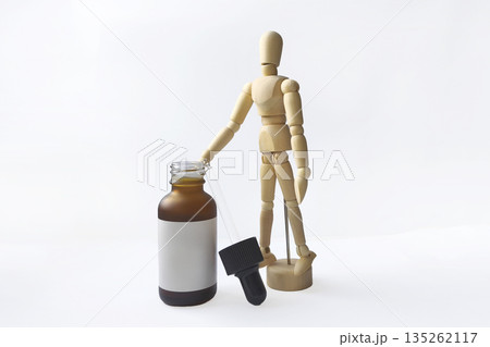 Wooden Dummy Doll stands next to a brown glass bottle with a white blank label and a pipette on a white background. 135262117