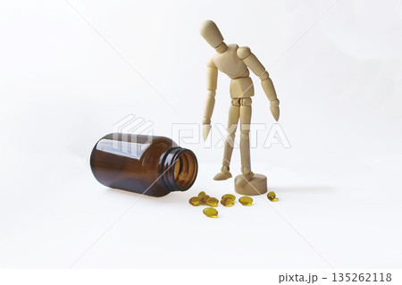 A wooden mannequin stands beside a tipped amber jar. Yellow capsules are scattered on a white surface. The scene suggests themes of health and wellness. A wooden mannequin stands beside a tipped amber jar. Yellow capsules are scattered on a white surface. The scene suggests themes of health and wellness. 135262118