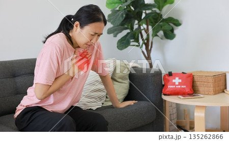 Asian woman sitting on sofa clutching chest with heart pain concept, healthcare, cardiology, emergency symptom, stress, medicine and heart attack awareness with ECG graphic overlay. 135262866