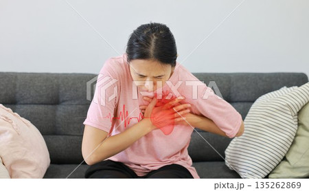 Asian woman sitting on sofa clutching chest with heart pain concept, healthcare, cardiology, emergency symptom, stress, medicine and heart attack awareness with ECG graphic overlay. 135262869